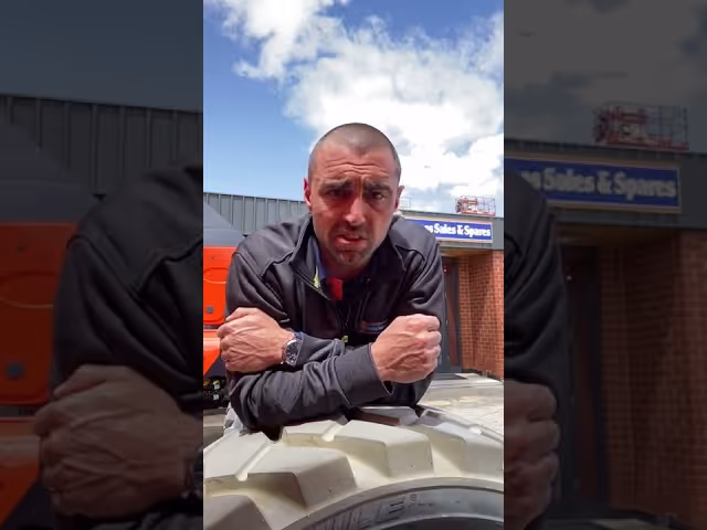 Man leaning on a large tire, looking directly at the camera, with a building in the background.