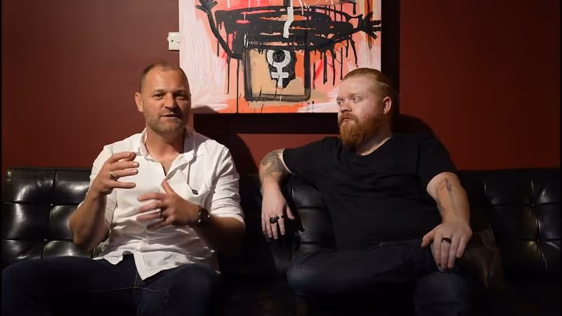 Two men sit on a black couch, talking animatedly against a dark red wall with artwork.