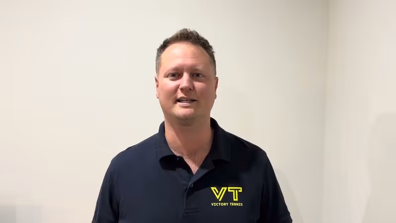 Man wearing a navy blue Victory Tennis polo shirt stands against a plain light-colored wall.