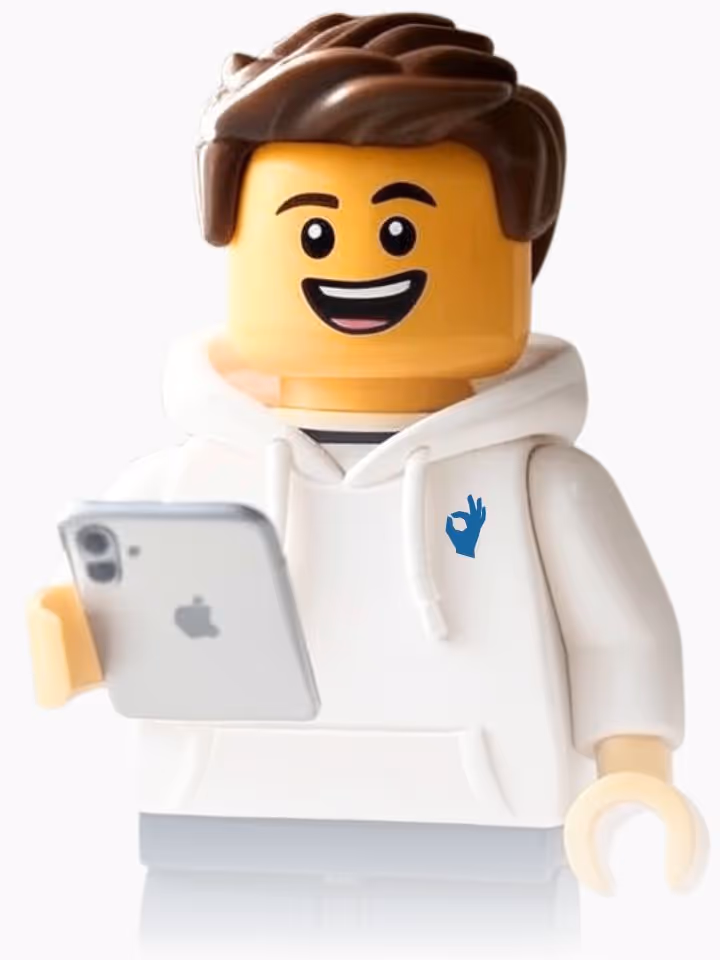 Smiling LEGO minifigure with brown hair wearing a white hoodie with a blue OK hand symbol, holding a silver smartphone.